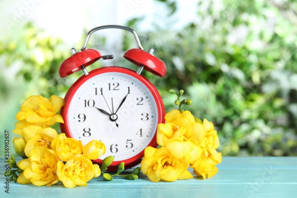 Fototapeta Red alarm clock and spring flowers on light blue wooden table against blurred greenery. Time change