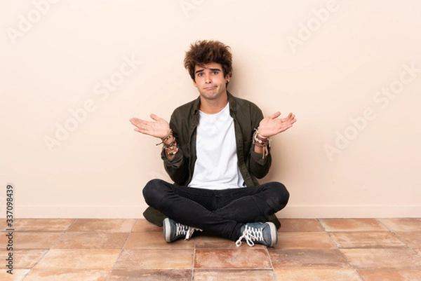 Fototapeta Young caucasian man sitting on the floor having doubts while raising hands