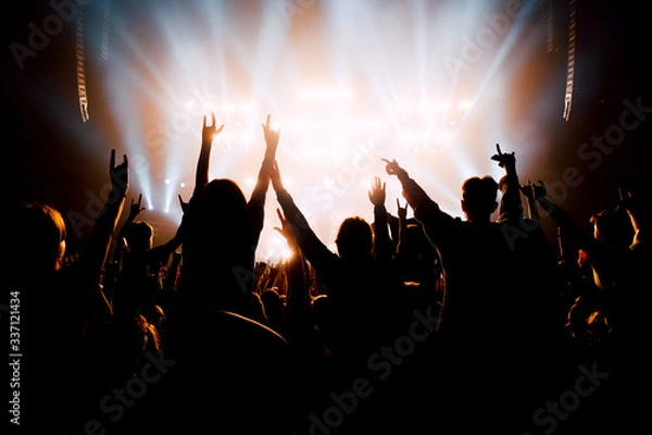 Fototapeta Crowd of people with raised up hands to the stage. Silhouettes in bright stage lights.