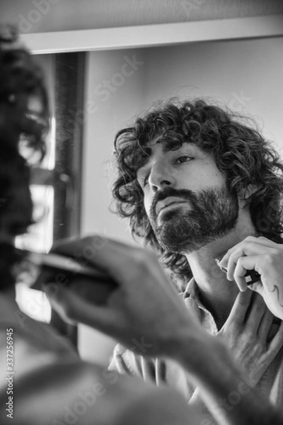 Fototapeta Handsome young man looking in the mirror while fixing his beard. Lifestyle