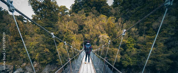 Obraz Hokitika Gorge, New Zealand, October 7, 2019: Beautiful panorama of a European man with a photographic backpack crossing a suspension bridge surrounded by leafy trees