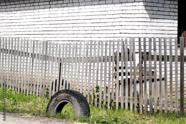 Fototapeta An old grey boardwalk wooden fence with parallel planks and a used tire on the green grass in front of a white brick wall on a bright sunny day. A concept of rural life with space for text