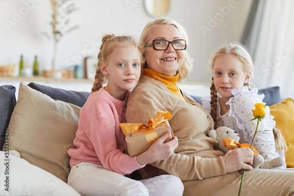 Fototapeta Portrait of happy grandmother in eyeglasses sitting on sofa together with her granddaughters and they smiling at camera