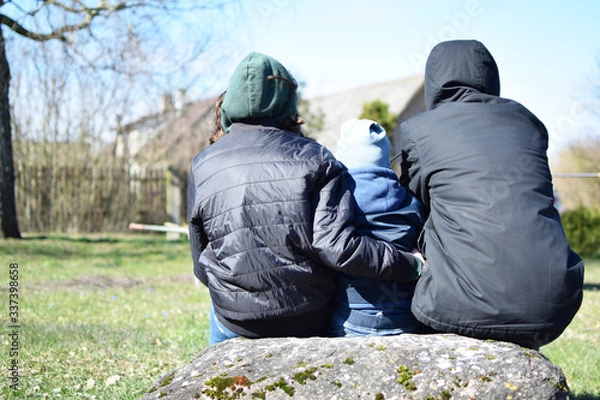 Obraz Teenage boy, girl and little child sitting on stone outdoors in backyard looking forward and talking about life in cold spring day, back view.
