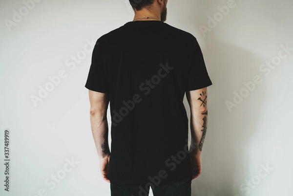 Fototapeta Young hipster wearing black jeans and blank black t-shirt with empty space for  logo, text or design. mock-up of t-shirt, white wall in the background. Back view.