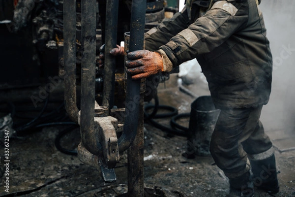 Fototapeta Work driller in red uniform, in helmet and goggles. He uses a hydraulic wrench to screw drill pipes to lower them into an oil well and continue drilling it. The concept of a working person.