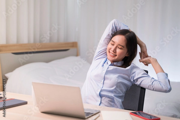 Fototapeta Attractive Asian woman works from home with laptop on desk in bedroom. Young girl raises arms, stretching to relieve pain from office syndrome. Shelter in place from COVID-19 and Stay Home concept.