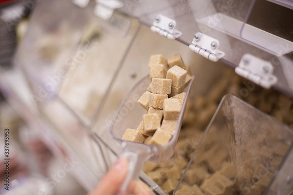 Fototapeta A woman buys brown sugar cubes in a grocery store. Girl puts cane sugar with a plastic scoop in the bulk department