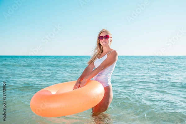 Fototapeta Girl with inflatable rubber enters the sea. Model goes into clear blue water on a hot day and holding orange ring.