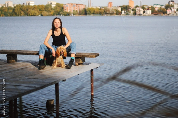 Fototapeta Girl with her dog, English Cocker Spaniel, sitting on the bench near the lake