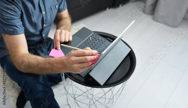 Fototapeta Young man in casual clothes sitting on floor and working on his laptop computer at home. Stay home and work online. Quarantine concept, panoramic banner