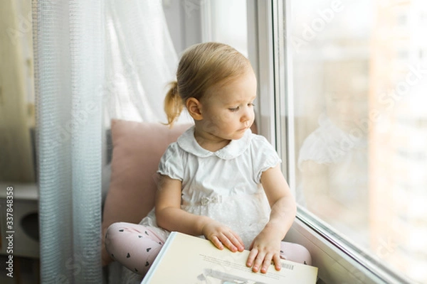 Fototapeta Little girl sitting on the windowsill of the house, self-isolation at home in 2020