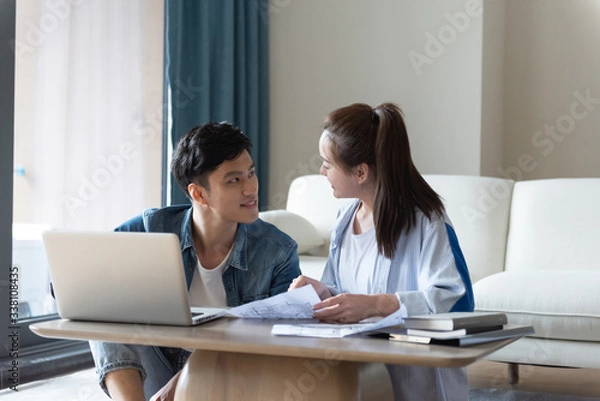 Fototapeta A young asian couple is sitting on a carpet using a computer