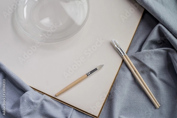 Obraz Creative space: cristal jar, brushes and blank paper on blue fabric.