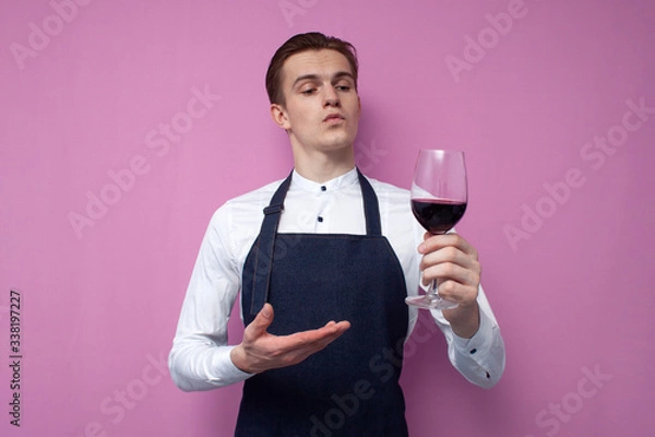 Obraz young sommelier guy in a white shirt and apron holds a glass of red wine and appreciates the taste, an expert tastes wine on a colored background