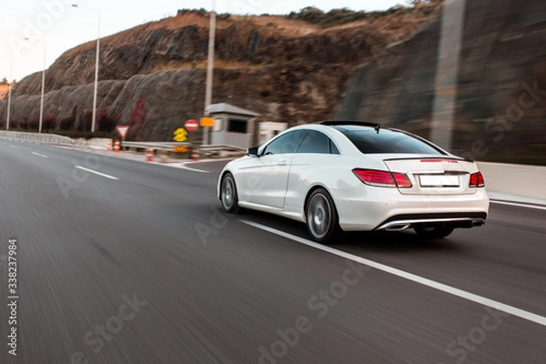 Obraz White business class sedan car driving on the highway across green hills