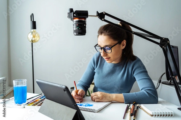 Fototapeta Online content creator vlogger. A young women blogger with glasses removes the content for the blog, training courses. The concept of online learning to draw on walercolor.