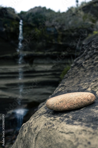 Fototapeta 
Image of a stone resting on the hillside with the waterfall in the background taken on a cloudy day at Truman Track, New Zealand