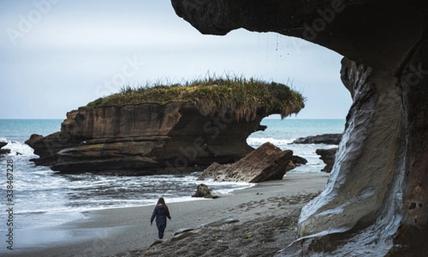 Obraz Truman Track, Nowa Zelandia, 8 października 2019 r.: Piękny obraz blondynki europejskiej spacerującej po plaży z formacją skalną w środku zabranego w pochmurny zimowy dzień