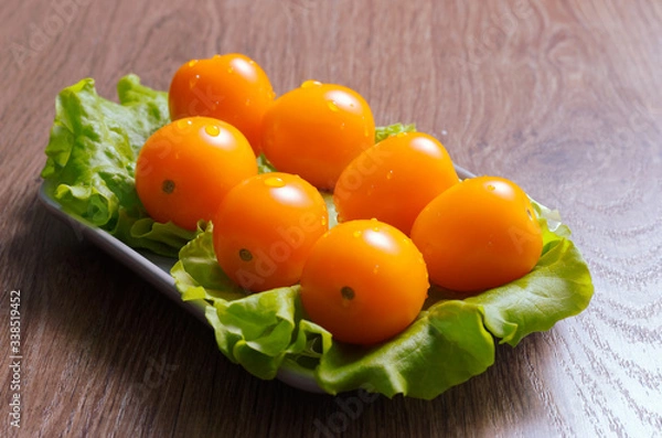 Obraz Yellow cherry tomatoes covered with drops of water on a lettuce leaf lie on a plate. Plate is on a wooden tabletop.
