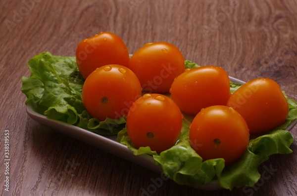 Obraz Yellow cherry tomatoes covered with drops of water on a lettuce leaf lie on a plate. Plate is on a wooden tabletop.