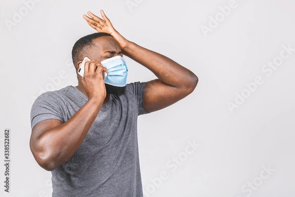 Fototapeta Shocked young african american man in sterile face mask posing isolated on white wall background. Epidemic pandemic coronavirus 2019-ncov sars covid-19 flu virus concept. Using phone.