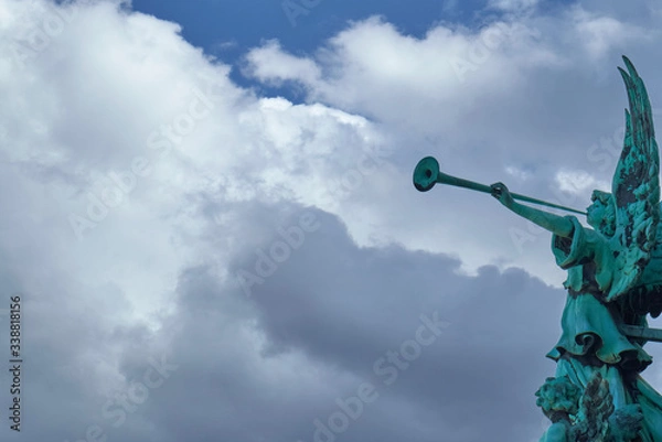 Fototapeta Trumpeting old bronze angel statue covered with green verdigris playing a celestial fanfare against a sunny sky with dramatic clouds.