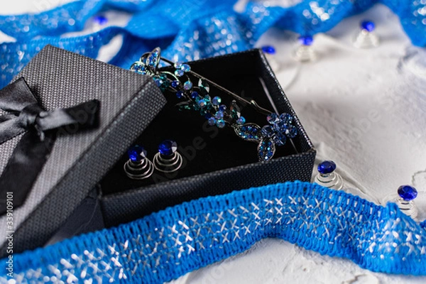 Fototapeta Beautiful brooch with blue and aquamarine stones and rhinestones of different sizes, metal base pins in a gift box with a black bow, around the ribbon with silver threads and springs.