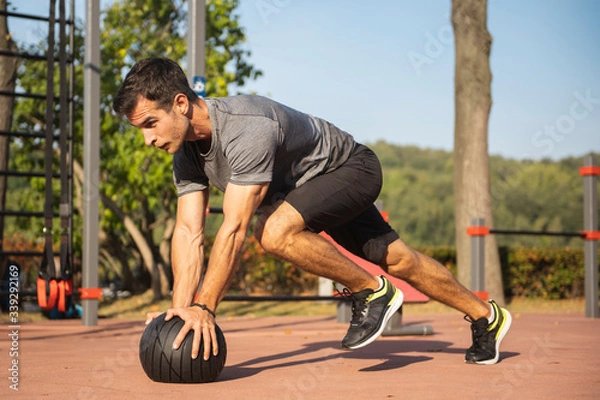 Obraz Fit guy doing exercises using a ball outdoors. Young athletic man training in city park