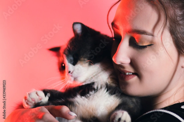 Fototapeta portrait of a young happy woman with a cat in her arms in red color lighting in the studio, girl plays with pet, lovely animals