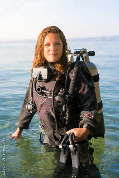 Fototapeta Female Scuba Diving Instructor Standing in Water Wearing a Dry Suit, a Twin Tank and Holding Fins