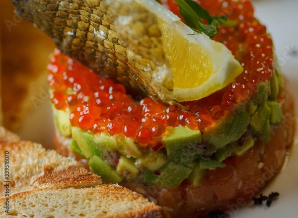Fototapeta Tartar with red caviar,avocado and fish .The tartar stands on a white plate and next to it lies butter with fried bread.Decorated of fish scale and lemon.