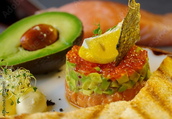 Fototapeta Tartar with red caviar,avocado and fish .The tartar stands on a white plate and next to it lies butter with fried bread.Decorated of fish scale and lemon.