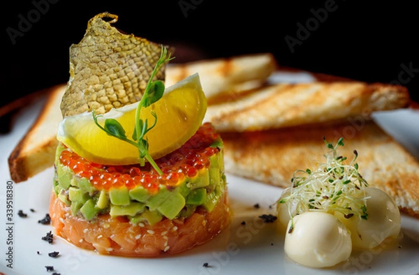 Fototapeta Tartar with red caviar,avocado and fish .The tartar stands on a white plate and next to it lies butter with fried bread.Decorated of fish scale and lemon.