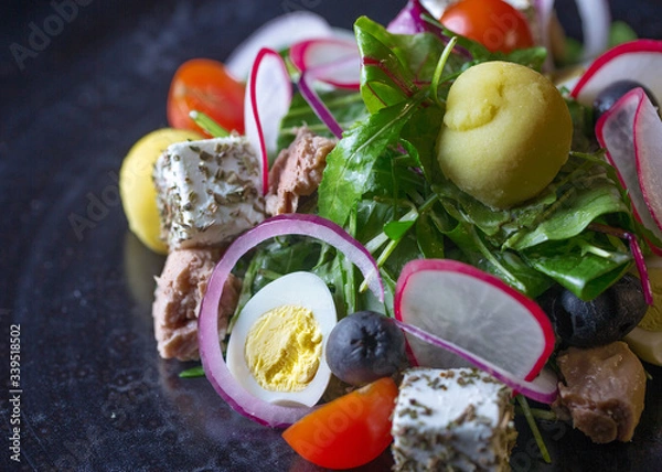 Fototapeta A salad of cherry tomatoes,arugula,lots of herbs,radishes,eggs,cheese and some meat.All the ingridients are on a dark table.