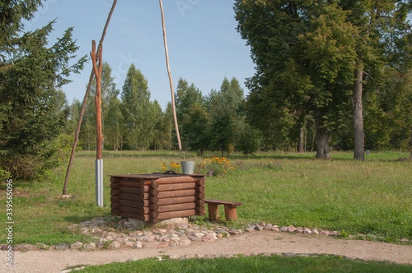 Fototapeta Old village well-crane with bucket for water in the field among the trees. Forest and blue sky.