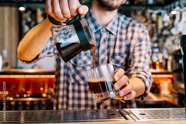 Fototapeta Alternative Coffee Brewing Method. 
Close-up of the hands of barista, pouring a black filter coffee from a teapot into a transparent glass on a bar counter in a cafe.