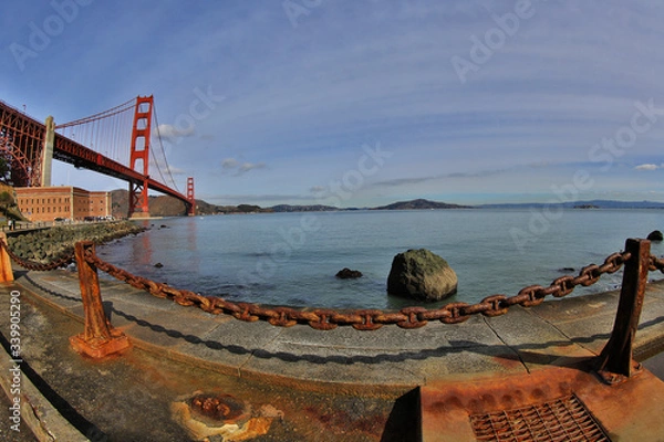Fototapeta Golden Gate Bridge with weathered harsh rusty chain in foreground contrasting calm natural bay in background.