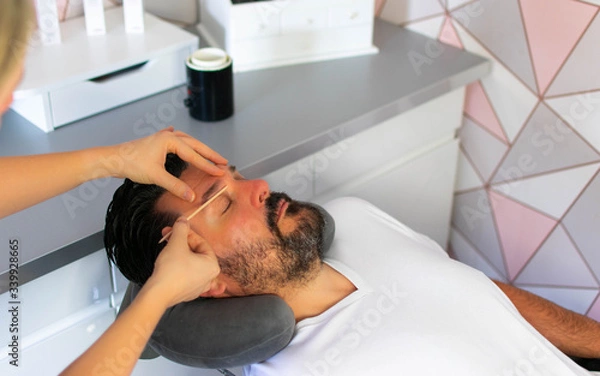 Fototapeta A beautician carries out an eyebrow wax on a relaxed man, removing unwanted hair and creating a flattering shape as part of a male grooming treatment and service in a fresh and modern beauty salon