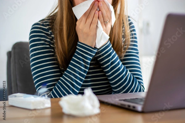 Fototapeta Sick woman working on laptop at home, covering nose and mouth when coughing and sneezing in tissue, ill freelance woman with cold. Coronavirus COVID-19 reducing of risk of spreading the infection.