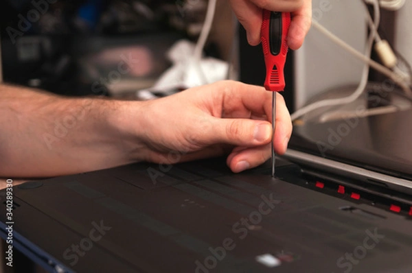 Fototapeta Male hands with screwdriver unscrew the back cover from laptop lying on table against a rack with different things