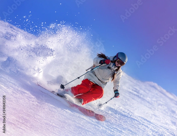 Obraz Girl On the Ski. a skier in a bright suit and outfit with long pigtails on her head rides on the track with swirls of fresh snow. Active winter holidays, skiing downhill in sunny day. Woman skier