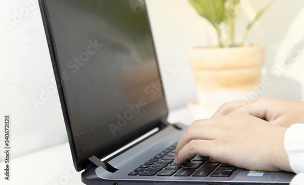 Fototapeta An Asian woman works from home using a single laptop on a white desk with a phone beside her.