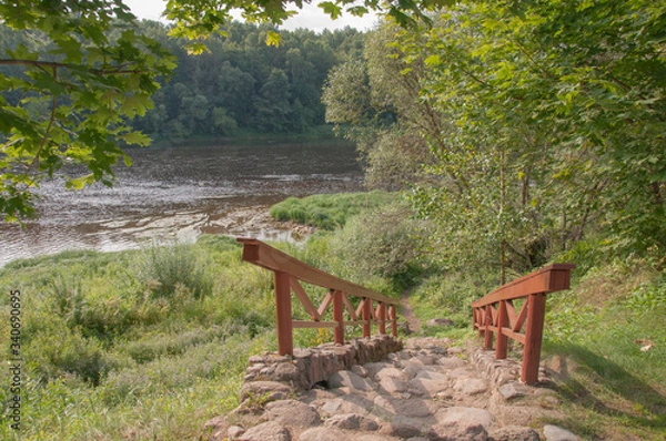 Fototapeta A stone staircase with wooden railings in the deciduous forest leading to a large river.