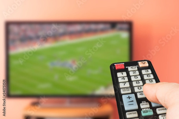 Fototapeta Caucasian man watching american football game on TV at home. Changing channels and adjusting volume with television remote control. 