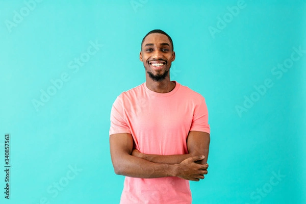 Obraz Portrait of a happy young man smiling with arms crossed, against blue studio background