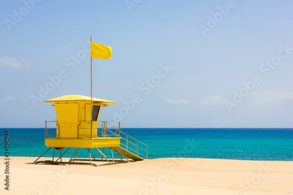 Fototapeta Colorful lifeguard tower off duty on empty beach in Corralejo natural park, Fuerteventura. Nobody on turquoise sea with yellow flag on top of baywatch station on sunny day. Summer vacation concept