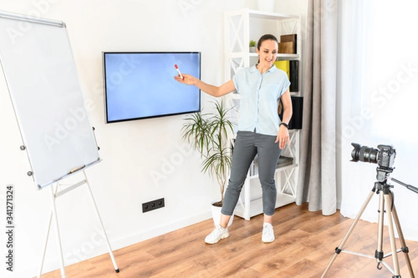Fototapeta A young woman is online teacher, tutor, mentor. A woman in smart casual is recording video classes, she is using flipchart and screen for explanation
