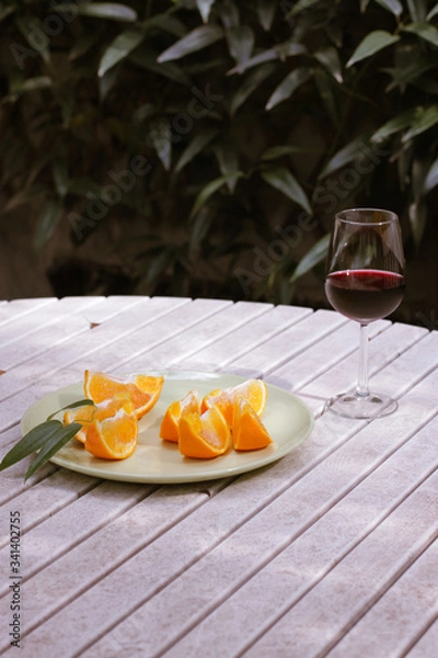 Obraz Red wine served outside in garden on white table with fresh orange in sun lights close up
