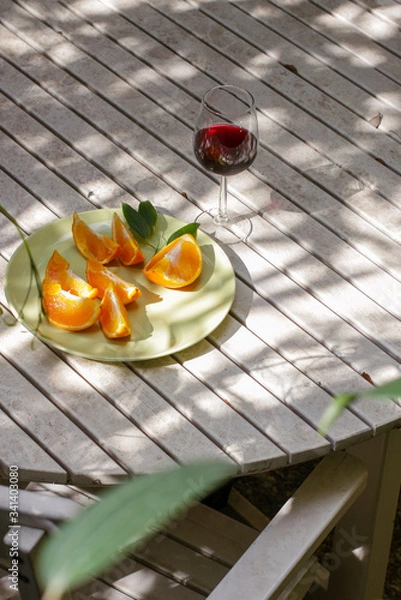 Obraz Red wine served outside in garden on white table with fresh orange in sun lights close up